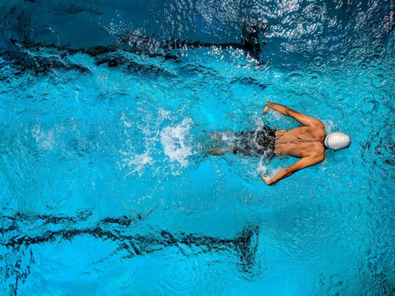 Home Top view of a swimmer wearing a cap, performing a front crawl stroke in a clear blue swimming pool.